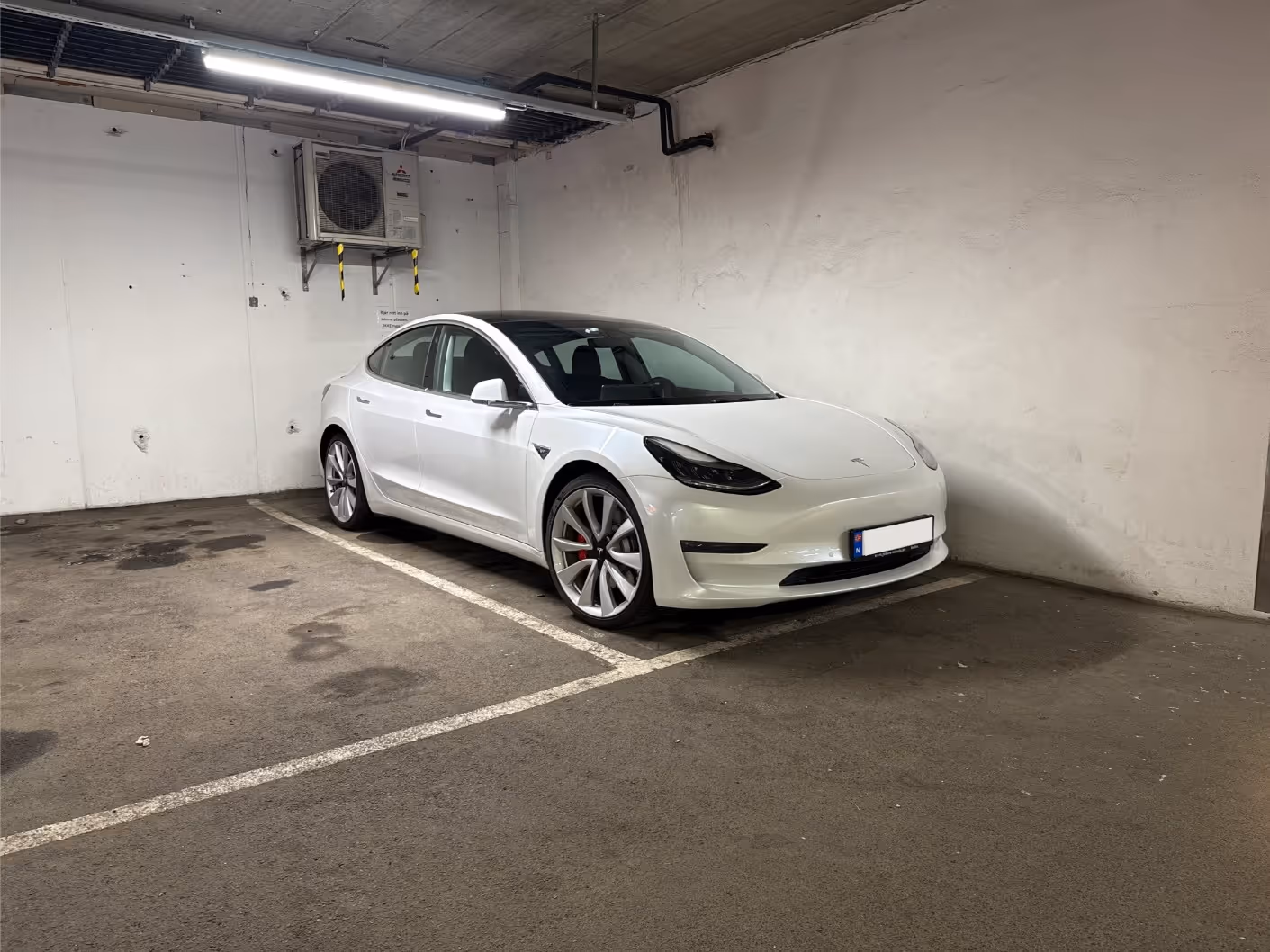 White Tesla Model 3 electric car parked in an indoor parking space with white walls and overhead lighting.