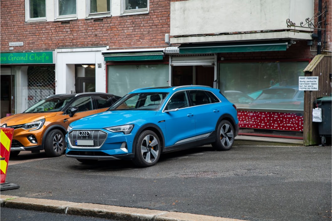 Blue Audi e-tron electric SUV parked next to an orange Renault Captur outside a brick building with shop windows.