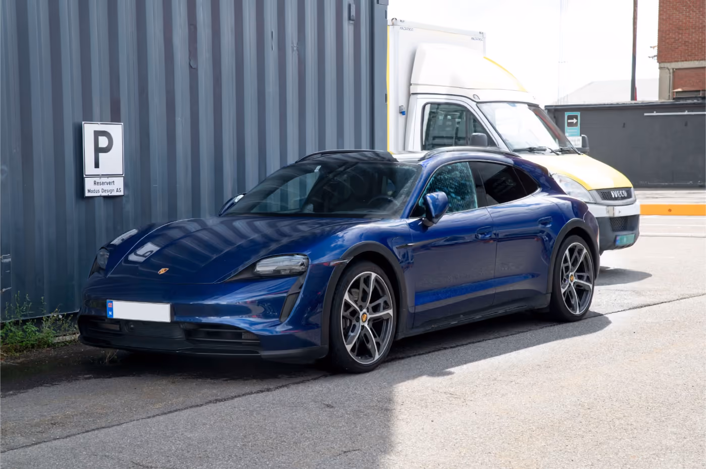 Blue Porsche Taycan electric car parked next to a grey corrugated wall and a white delivery van behind it.