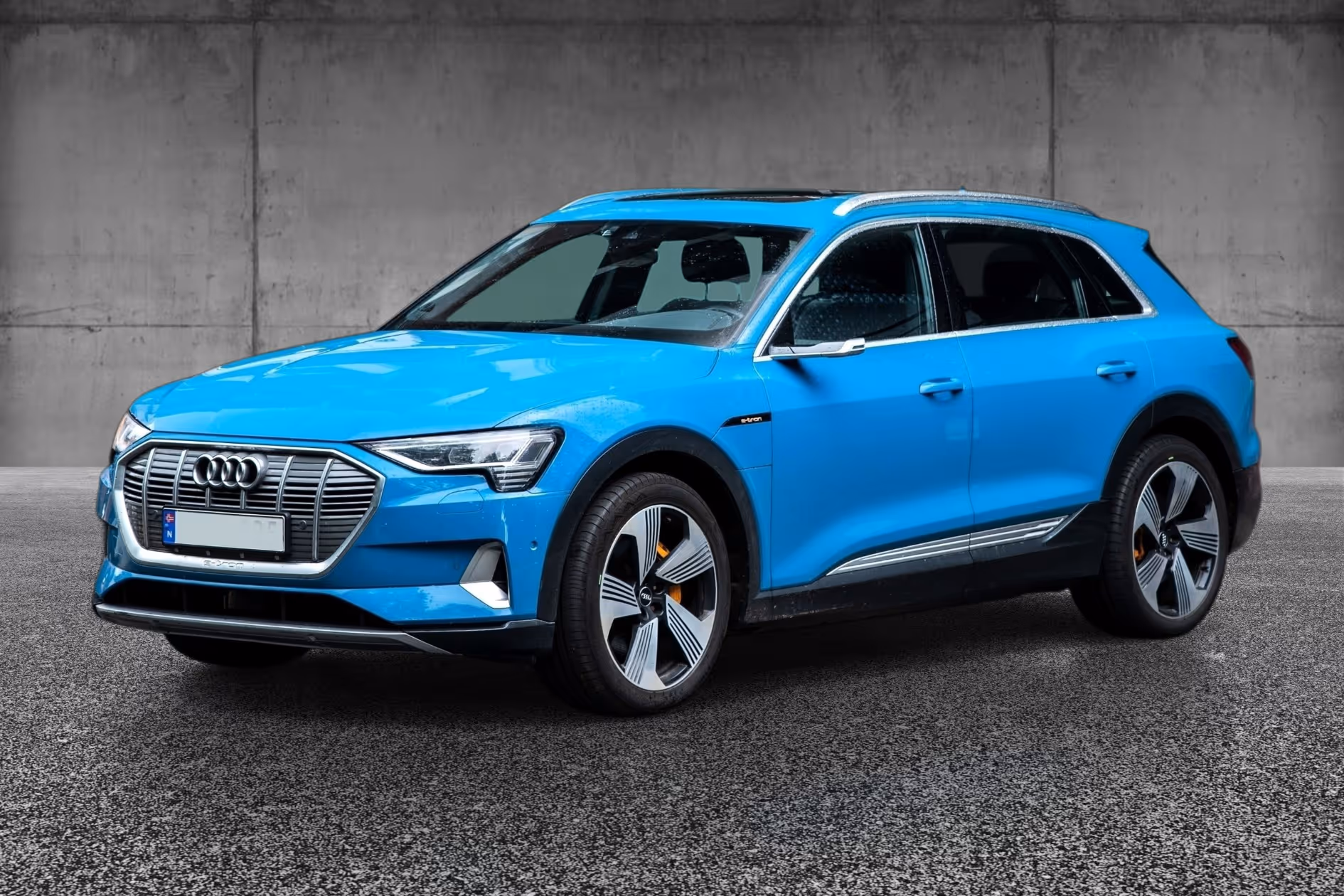 Blue Audi e-tron electric SUV parked on asphalt with a concrete wall background.
