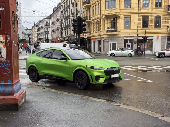Bright green Ford Mustang Mach-E electric SUV parked near a city intersection with historic buildings in the background.