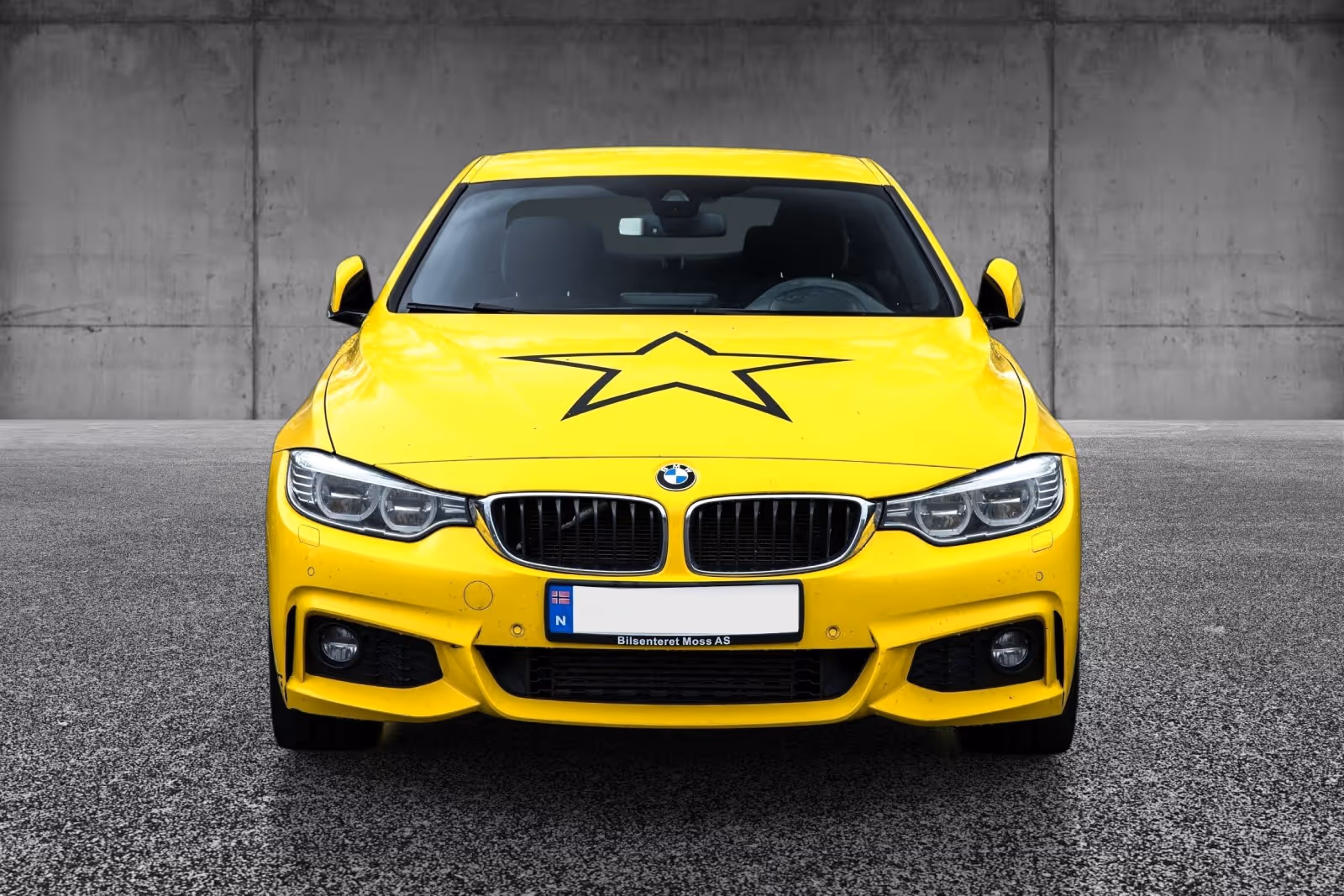 Front view of a bright yellow BMW car with a large black star decal on the hood parked on asphalt against a gray concrete wall.