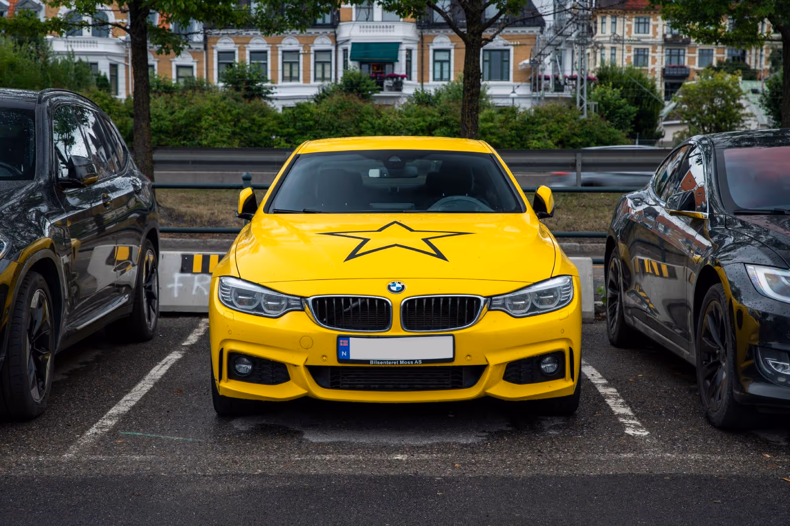 Yellow BMW car with a black star on its hood parked between two black cars in an urban parking lot.