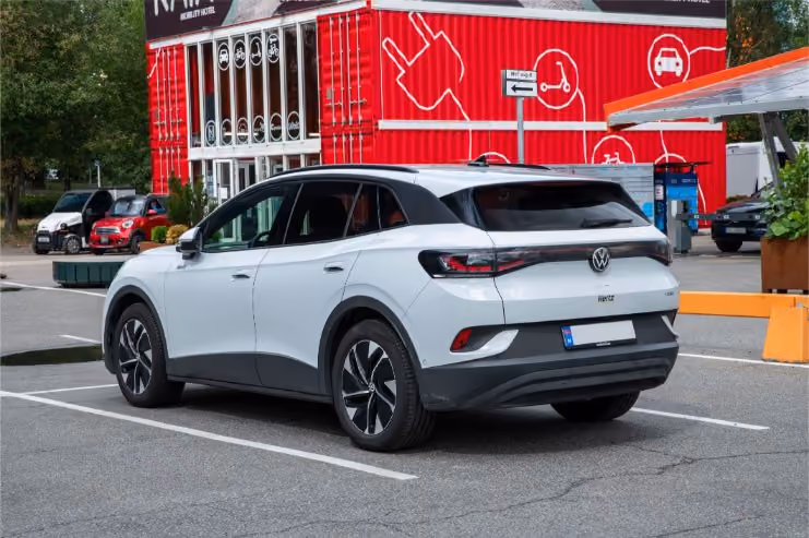 White Volkswagen ID.4 electric SUV parked in a lot near a red building with mobility and vehicle icons.