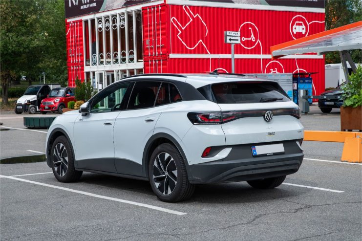 White Volkswagen ID.4 electric SUV parked in a lot near a red building with mobility and vehicle icons.