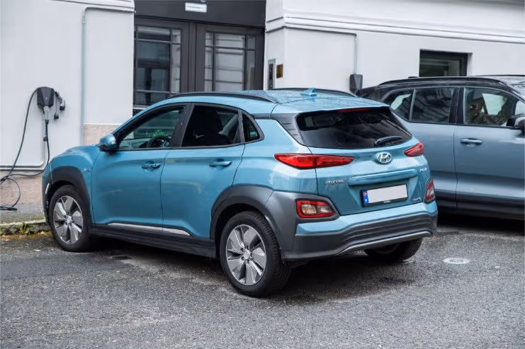 Light blue Hyundai Kona electric car parked near a charging station outside a building.