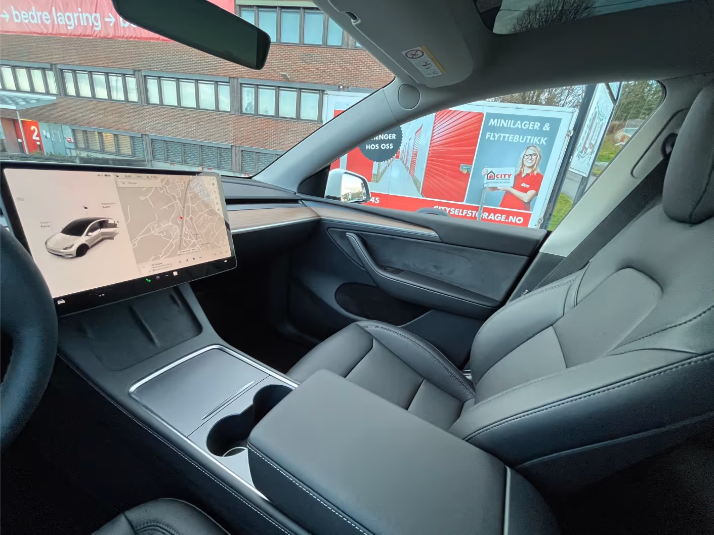 Interior of a modern car featuring a large touchscreen displaying a map and vehicle status, with black leather seats and a center console with cup holders.