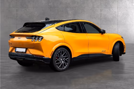 Rear-side view of a yellow Ford Mustang Mach-E GT electric SUV parked on gray concrete floor with a gray tiled wall background.