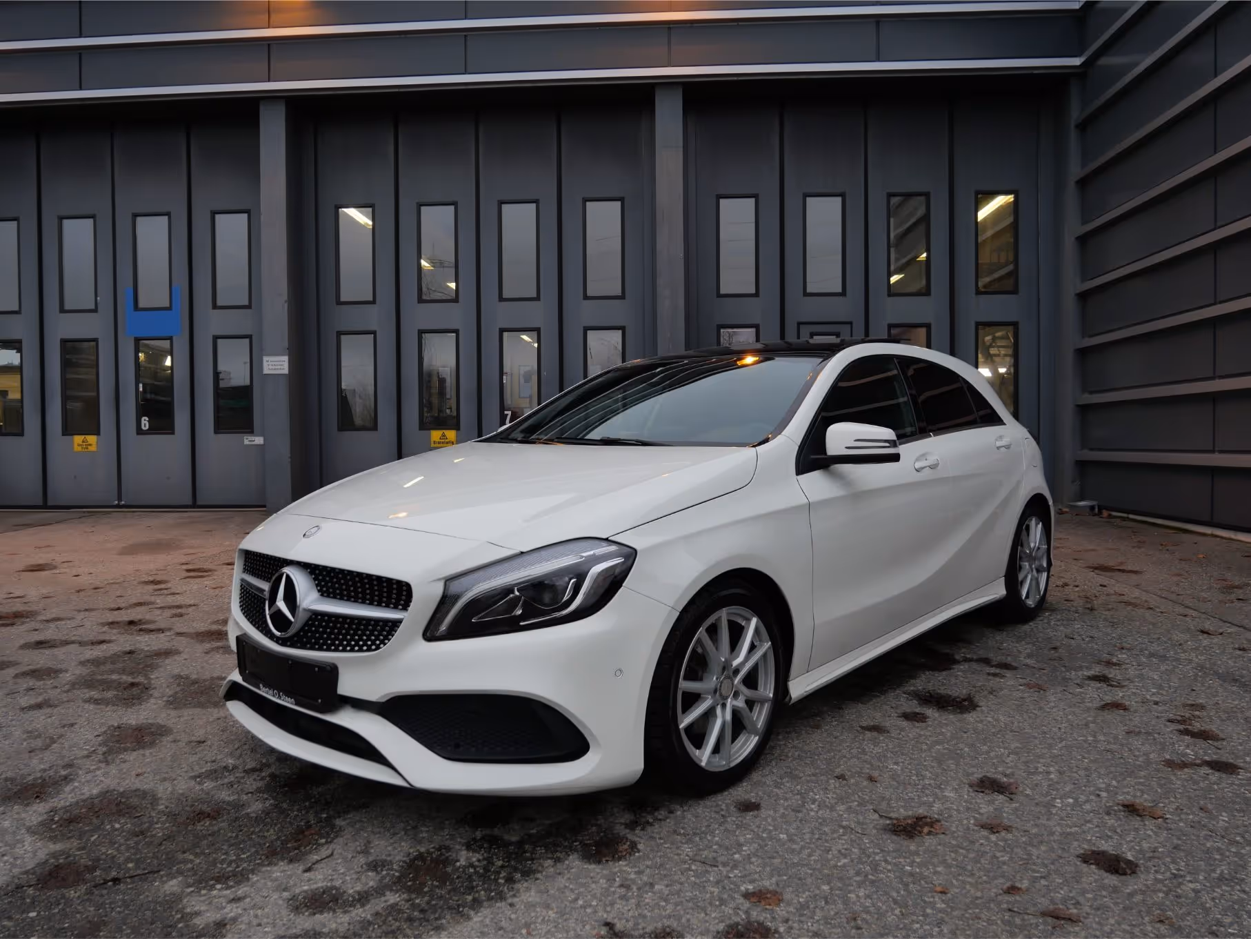 White Mercedes-Benz hatchback parked on a concrete surface in front of a dark building with multiple vertical windows.