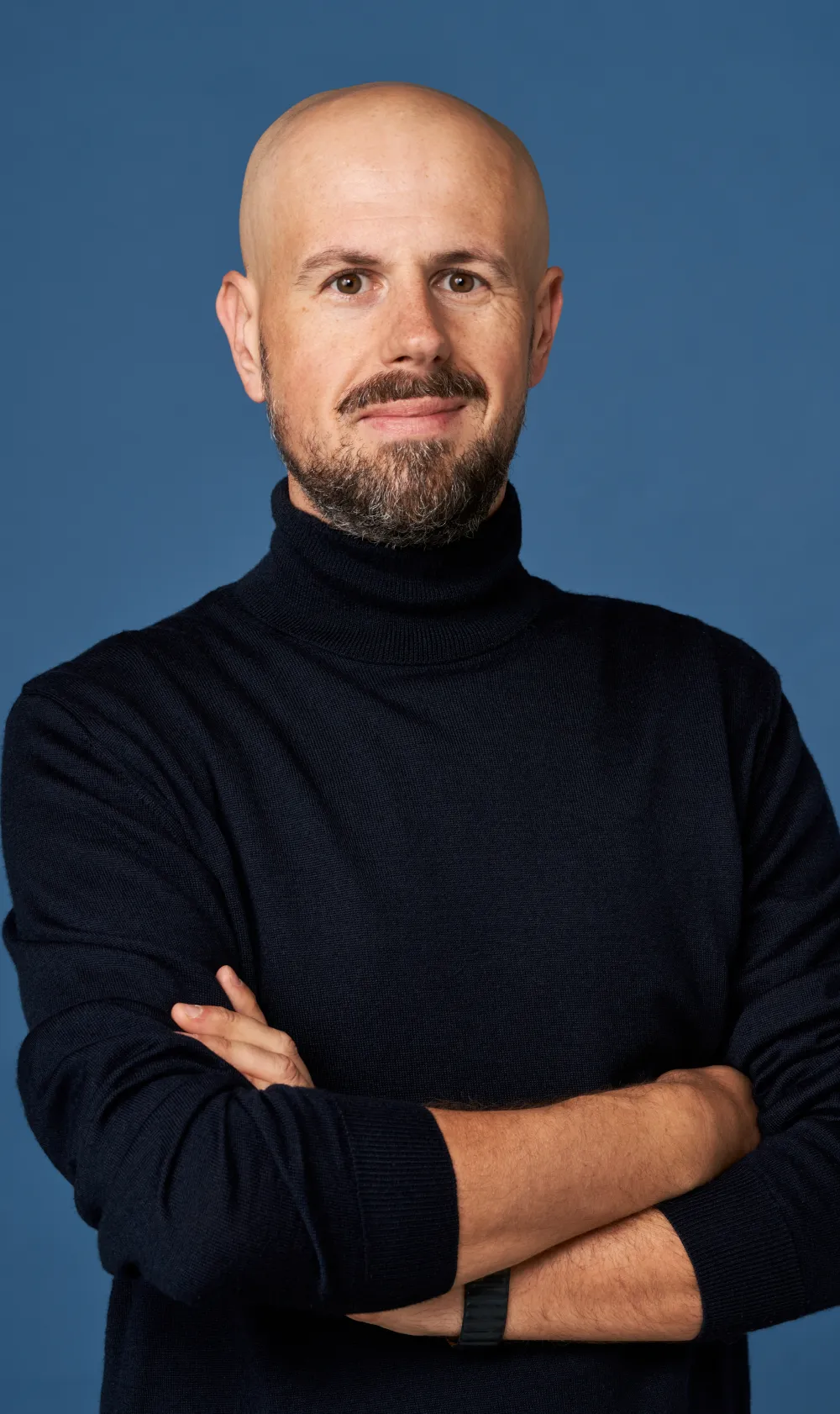 a business portrait of a man with beard and moustache, wearing black turtle neck with his arms crossed