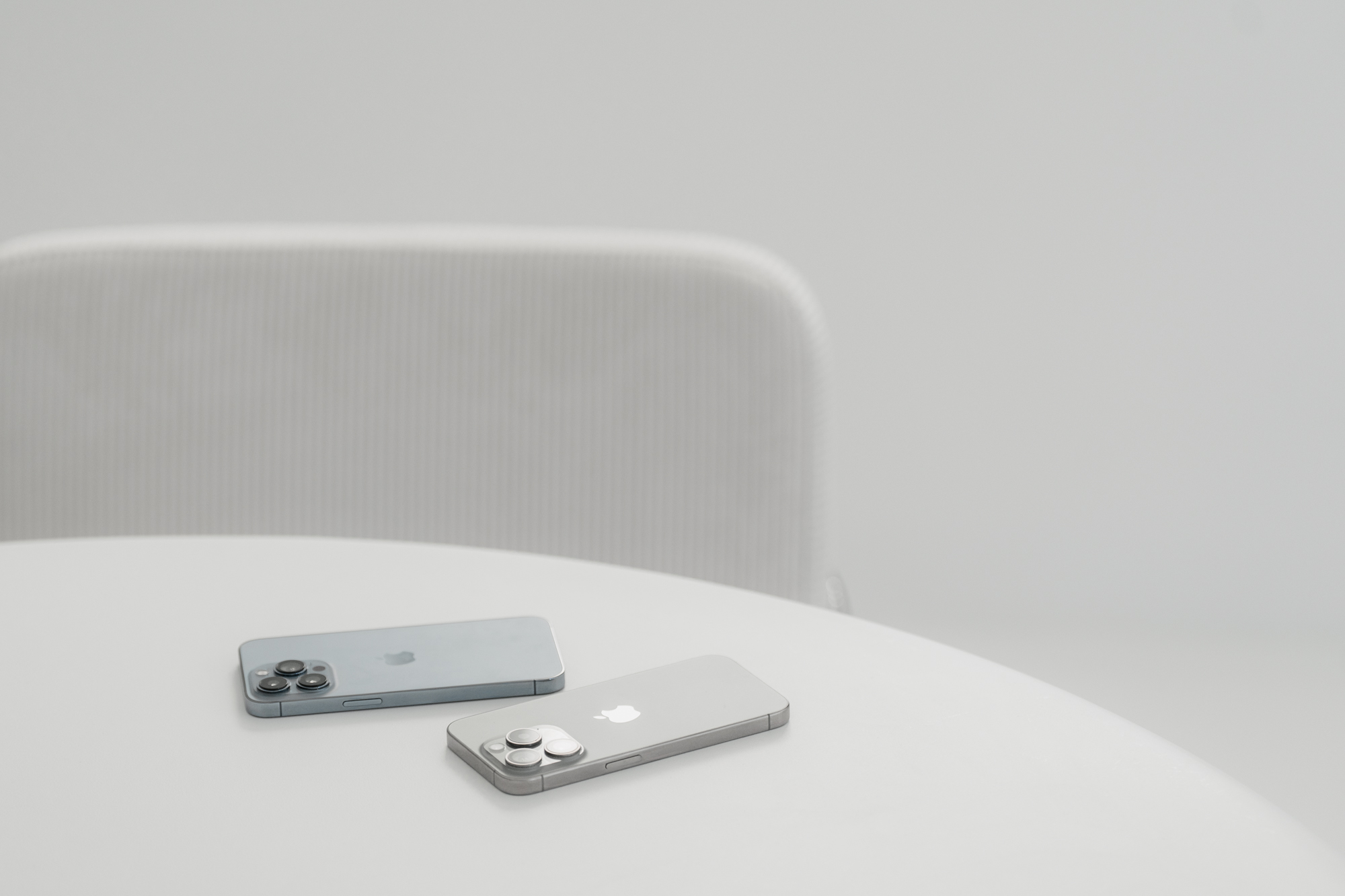 Zwei Apple iPhones mit Kameramodulen liegen auf einem weißen runden Tisch vor einem grauen Stuhl.