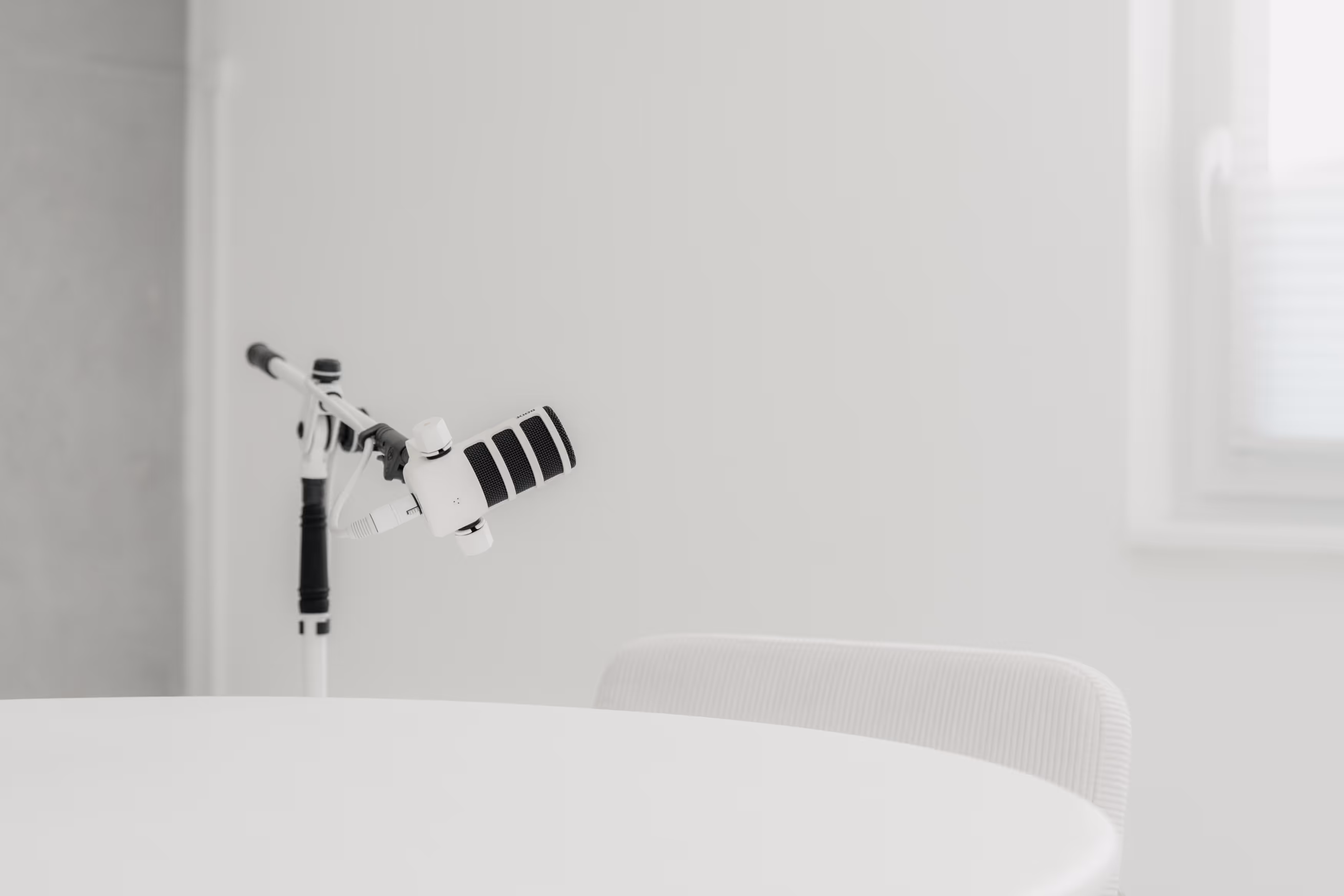 Weißes Podcast-Mikrofon mit schwarzem Schaumstoff, befestigt an einem Arm über einem weißen Tisch mit einem Stuhl im Hintergrund.