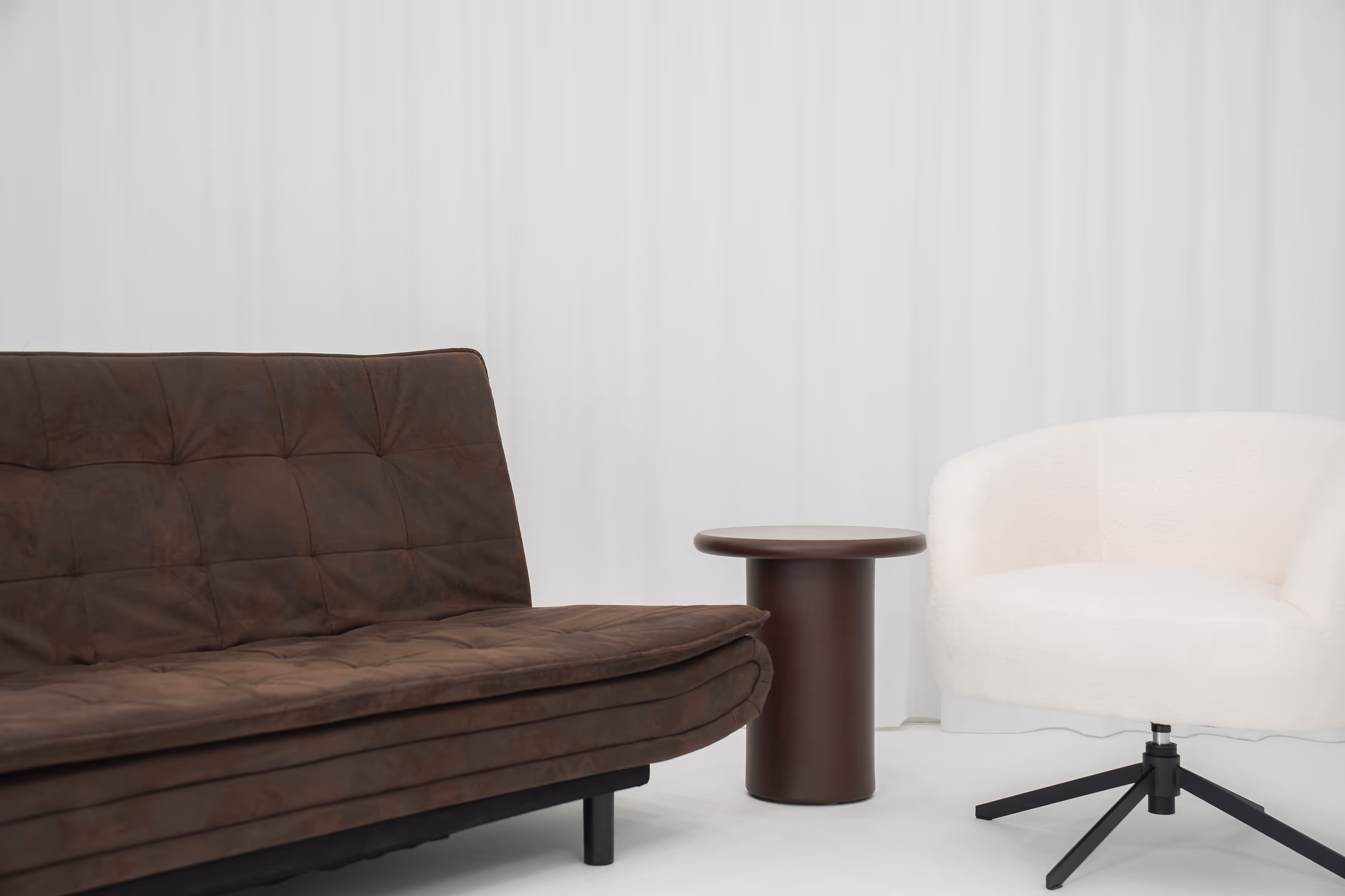 Braunes gepolstertes Sofa neben einem runden braunen Beistelltisch und einem weißen, drehbaren Sessel vor weißem Vorhang.