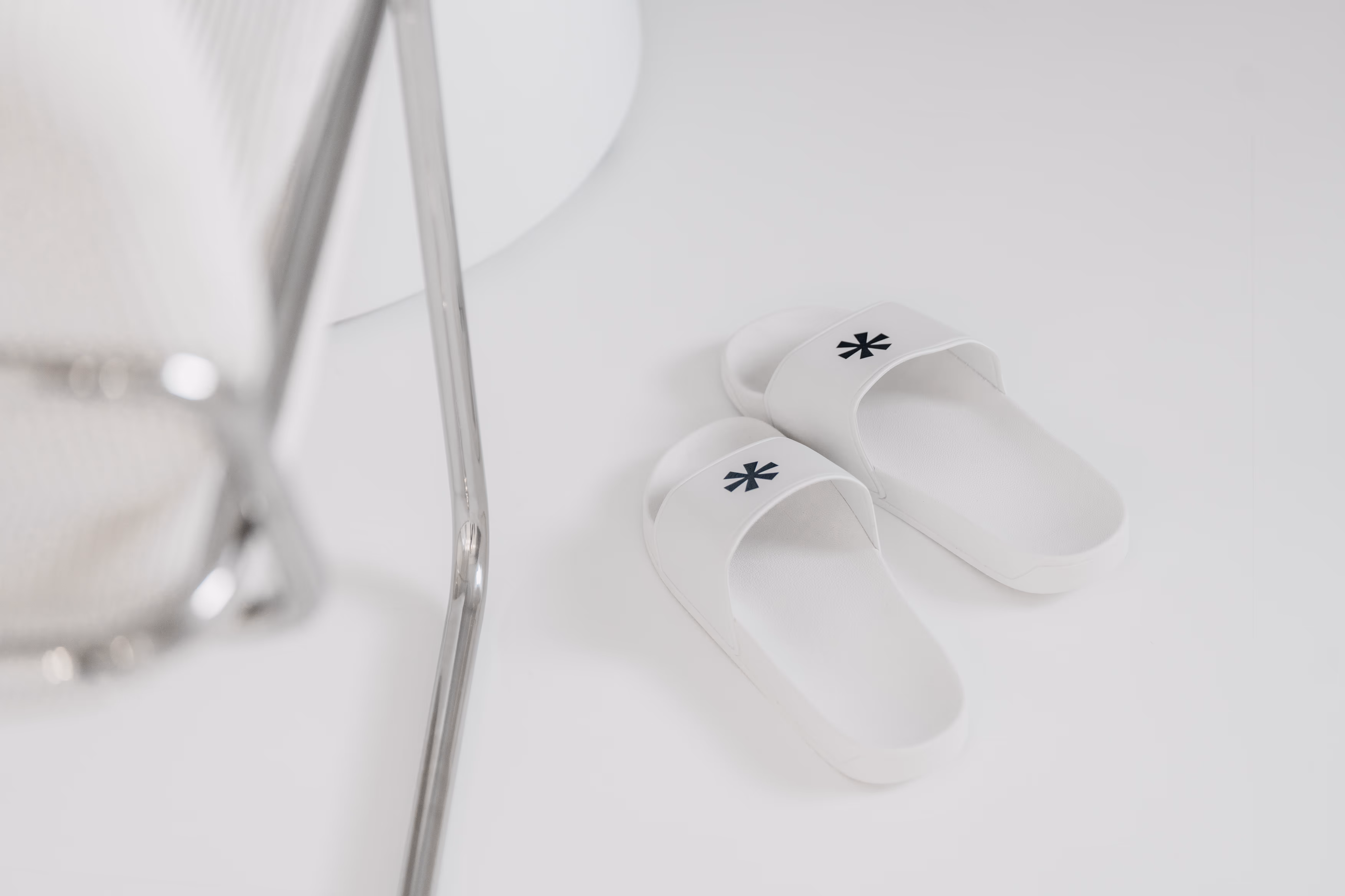 Ein Paar weiße Pantoffeln mit einem schwarzen Sternlogo auf einem weißen Boden neben einem Stuhl mit Metallrahmen.