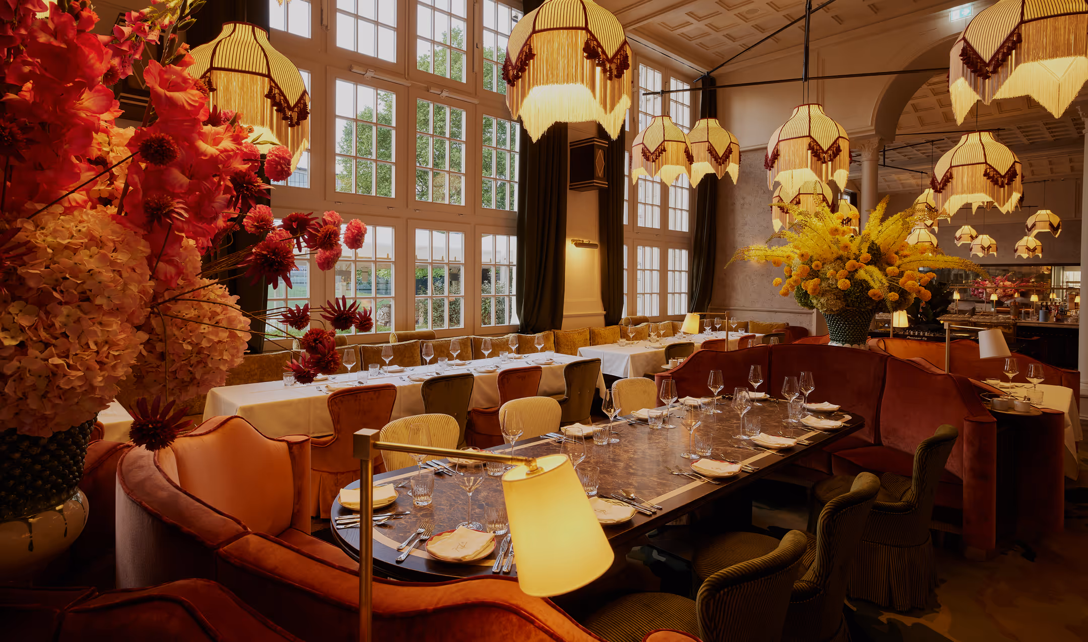Stilvoll gedeckter Gastraum im Pellicano Restaurant Hamburg mit großen Fenstern, warmem Licht, rotem und gelbem Blumenschmuck und elegantem Mobiliar.