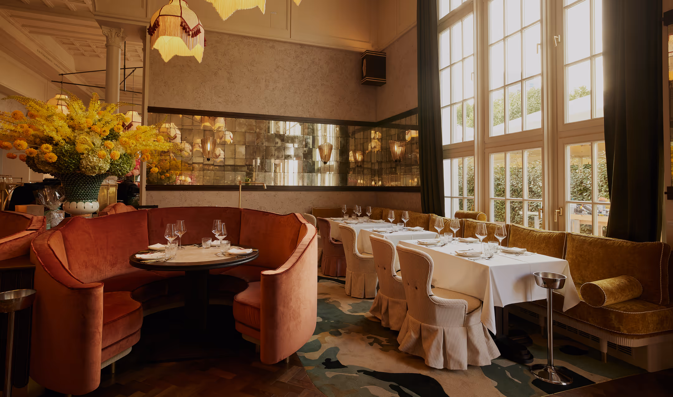 Elegant gedeckter Fensterbereich im Pellicano Restaurant Hamburg mit Spiegelwand, floraler Dekoration und samtbezogenen Sitzmöbeln in warmem Licht.