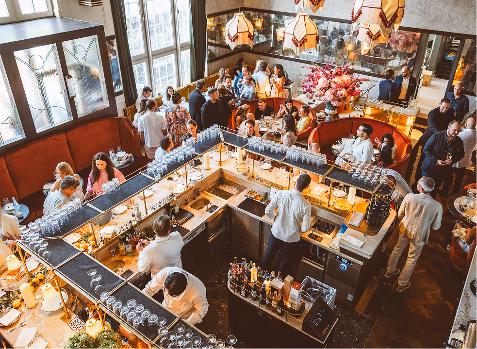 Lebendige Szene im Pellicano Restaurant Hamburg mit voll besetzter Bar, elegant gekleideten Gästen und stilvollem Interieur in warmer Tageslichtstimmung.