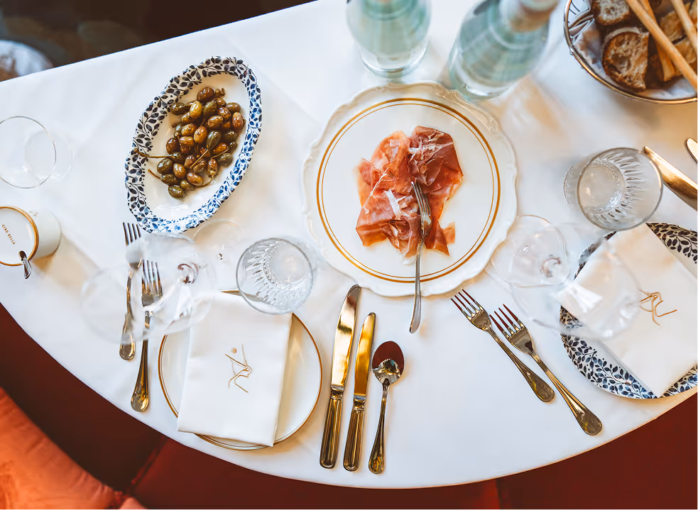 Elegant gedeckter Tisch im Pellicano Restaurant Hamburg mit Prosciutto, Oliven, Brot, Mineralwasser und edlem Besteck auf weißer Tischdecke.