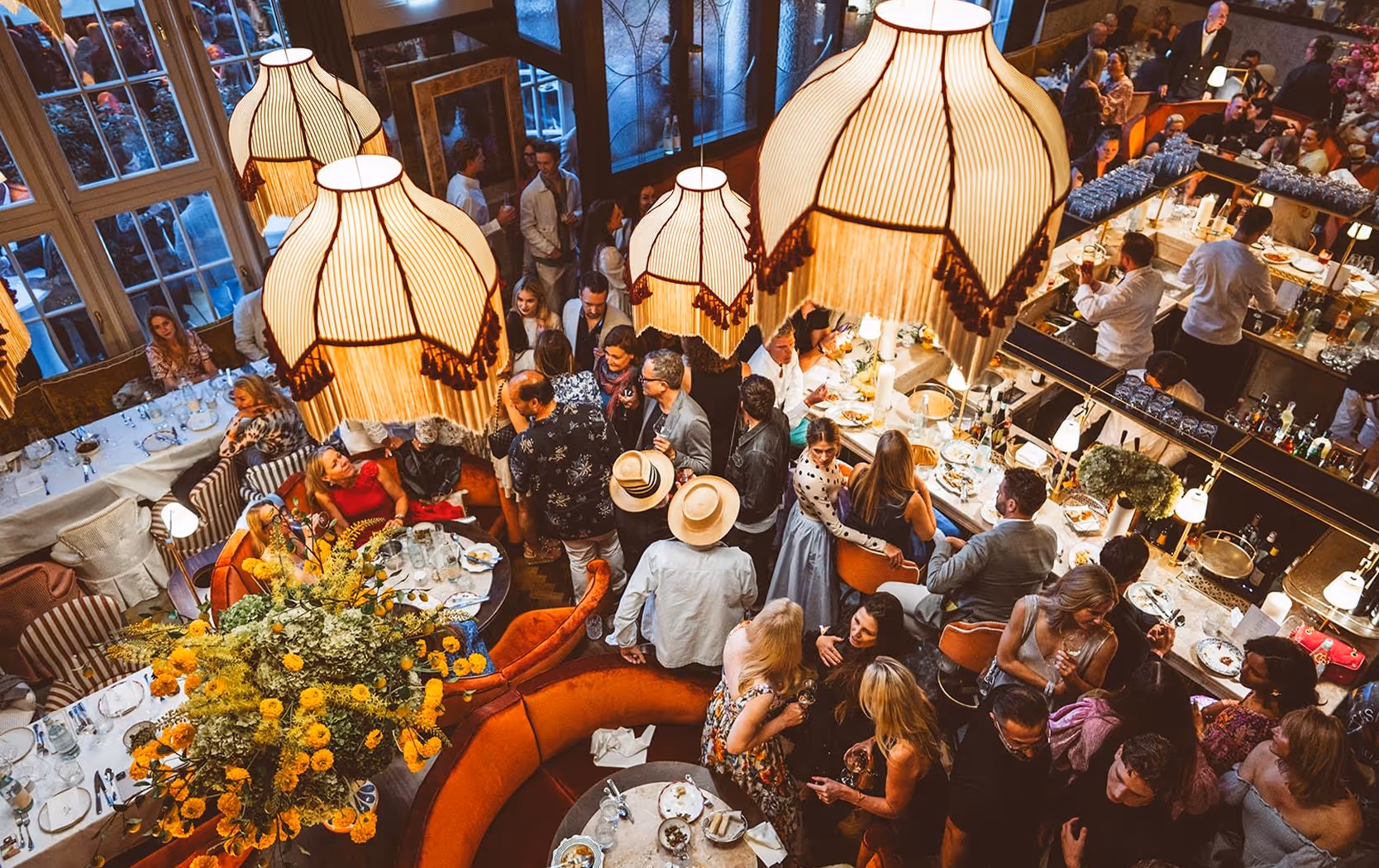 Abendliche Szene im Pellicano Restaurant Hamburg: Gäste in stilvoller Atmosphäre bei Drinks und Gesprächen, mit Blick auf die Bar und große florale Dekoration.