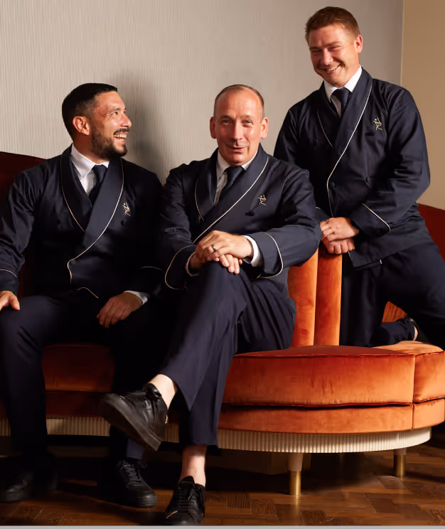Drei Mitarbeiter des Pellicano Restaurants Hamburg in stilvoller dunkler Uniform auf einem orangefarbenen Samtsofa – entspanntes Teamfoto mit eleganter Ausstrahlung.