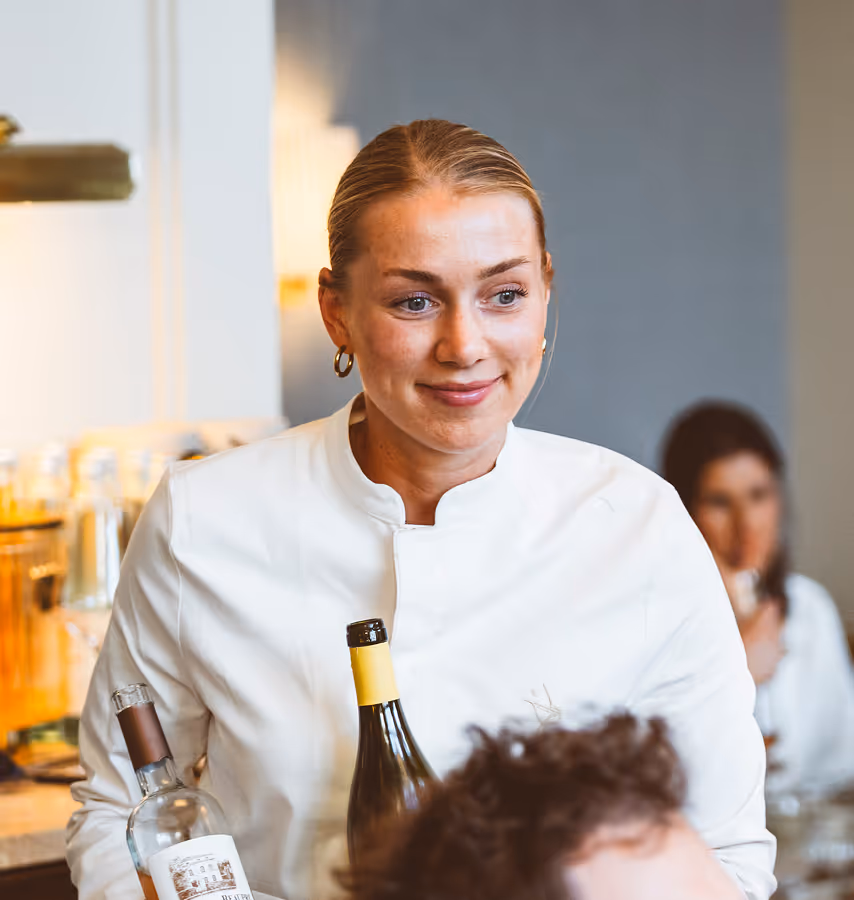 Freundliche Servicemitarbeiterin im weißen Outfit mit Weinflaschen in der Hand, lächelt während des Service im Pellicano Restaurant Hamburg.