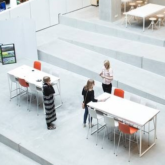 Gruppe set ovenfra i et stort lyst rum omgivet af enkelte borde, stole og skamler.