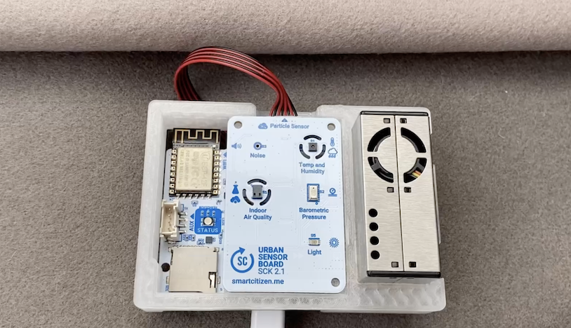 Sensorkit med en ledning og tekstbeskrivelse af sensorerne: Lys, lyd, luftkvalitet. Sensoren ligger på noget filt.