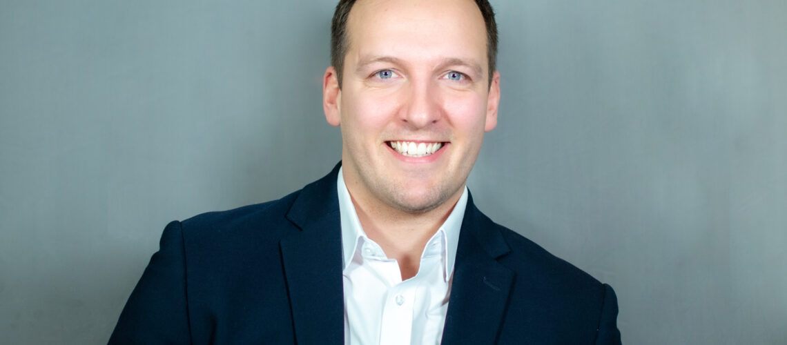 A man in a navy blazer and white shirt poses against a neutral gray background.