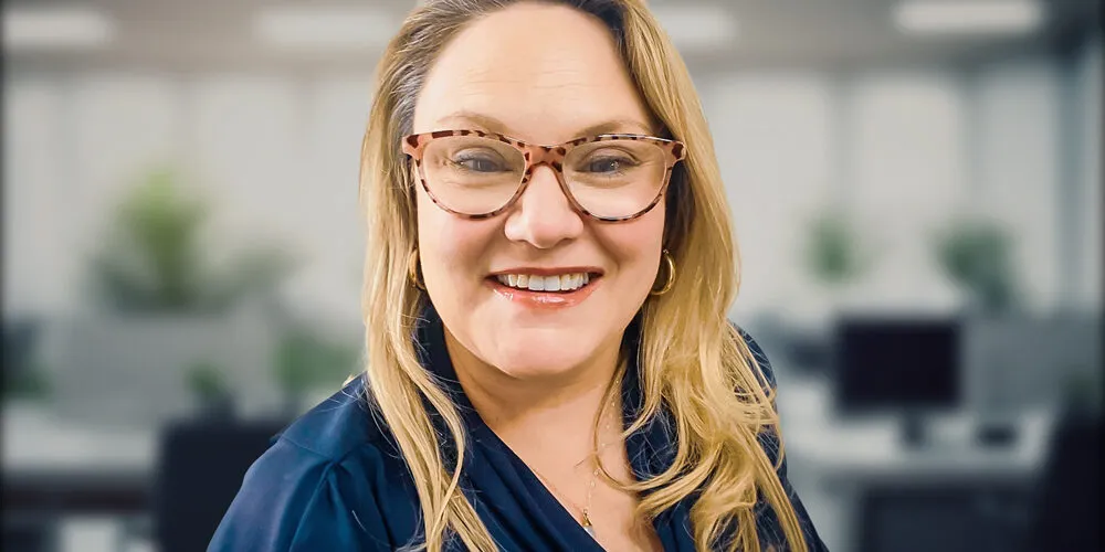 A woman with long blonde hair wearing a navy blouse stands in a modern office setting.
