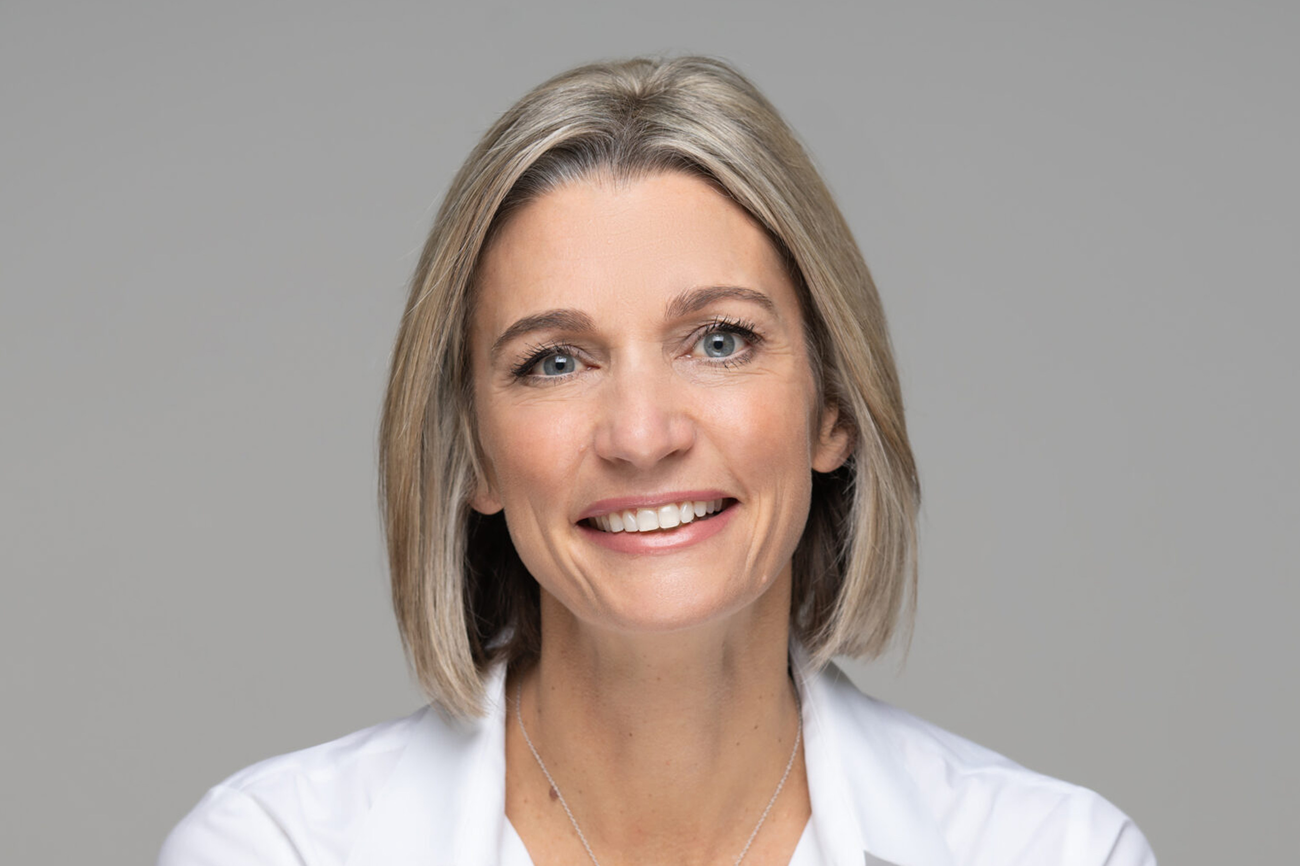 A woman with short, blonde hair wears a white shirt and a delicate necklace against a gray background.