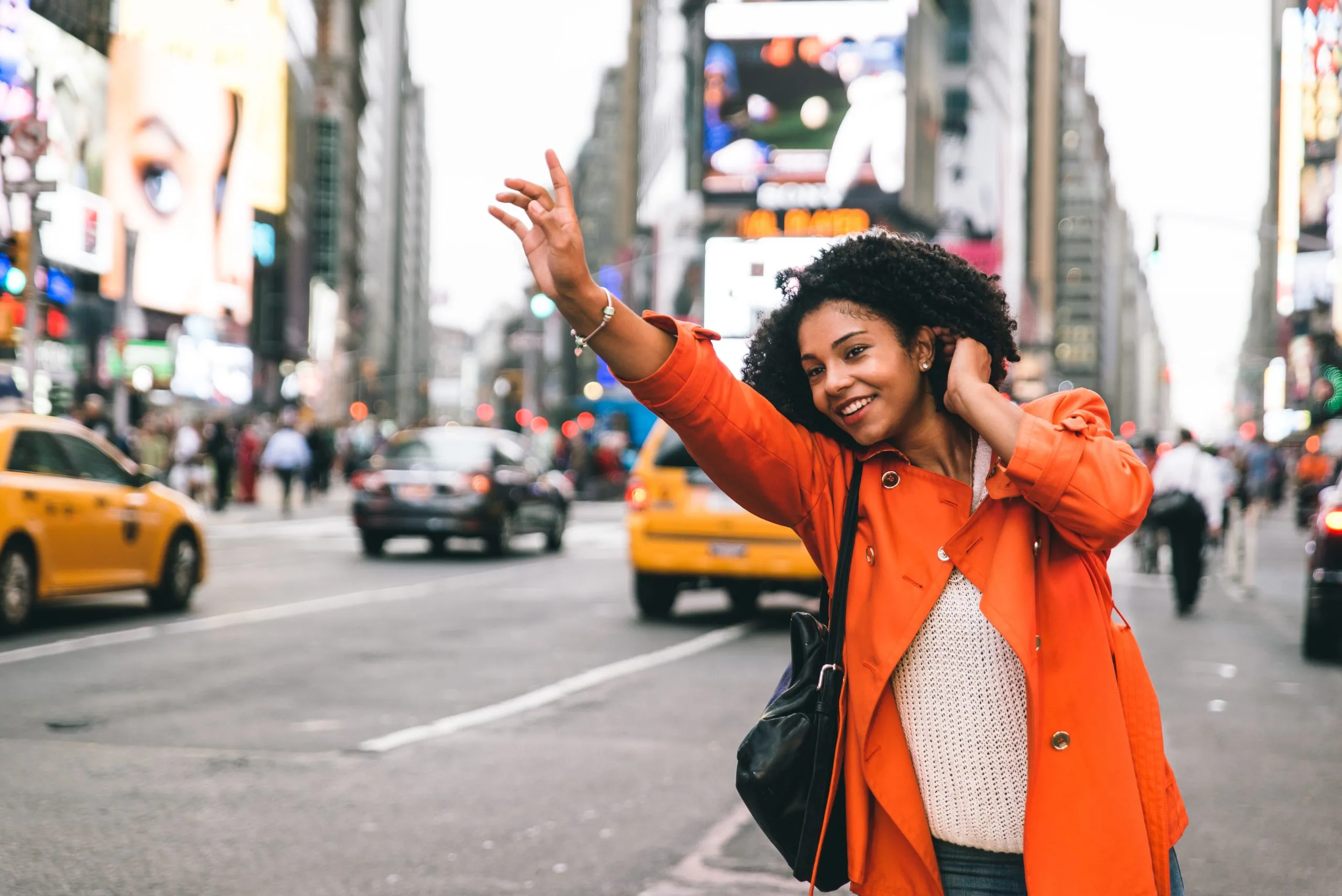 A woman in an orange coat hails a taxi on a busy city street with bright billboards and traffic.