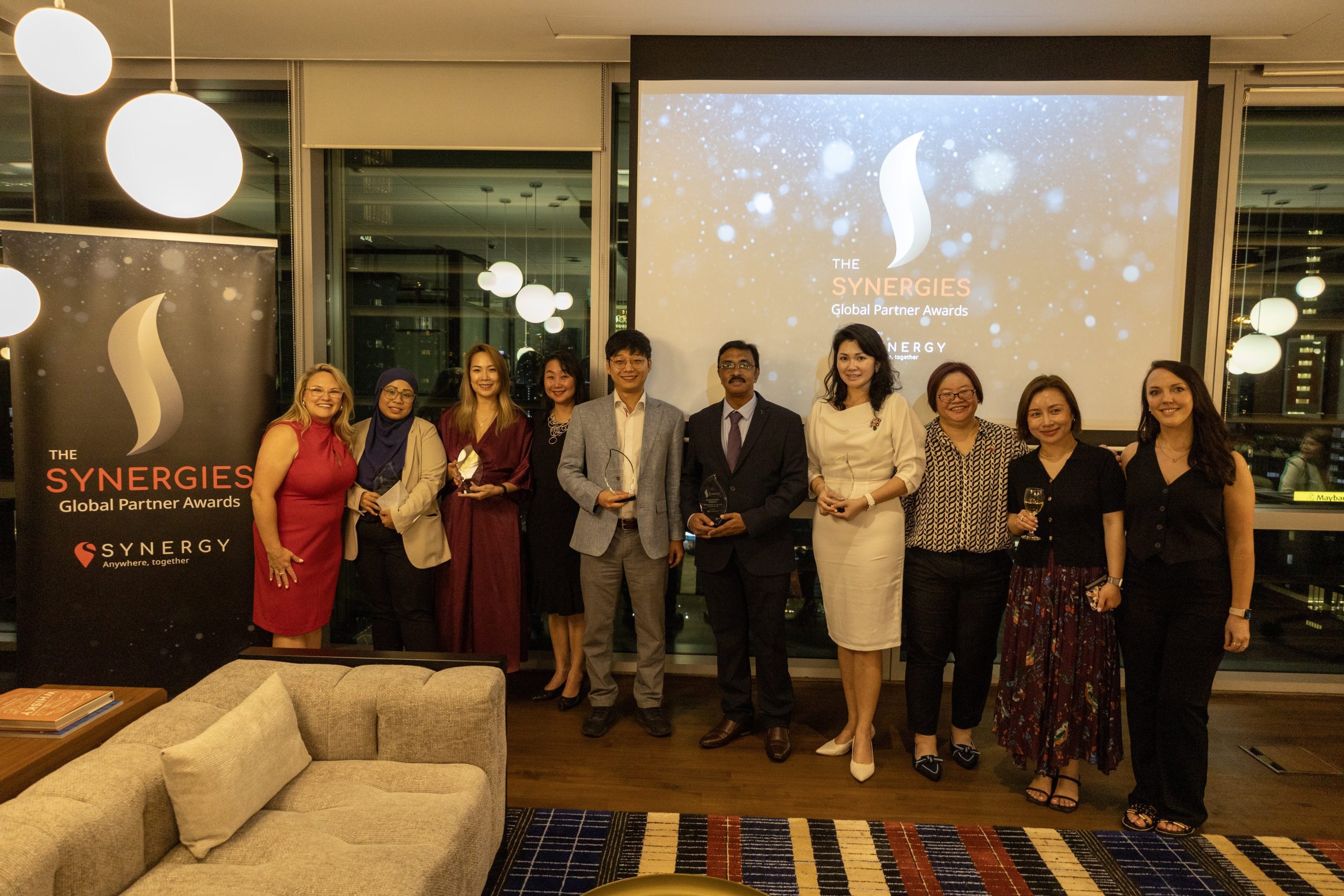A group of individuals pose at the Synergies Global Partner Awards event, holding trophies and awards.