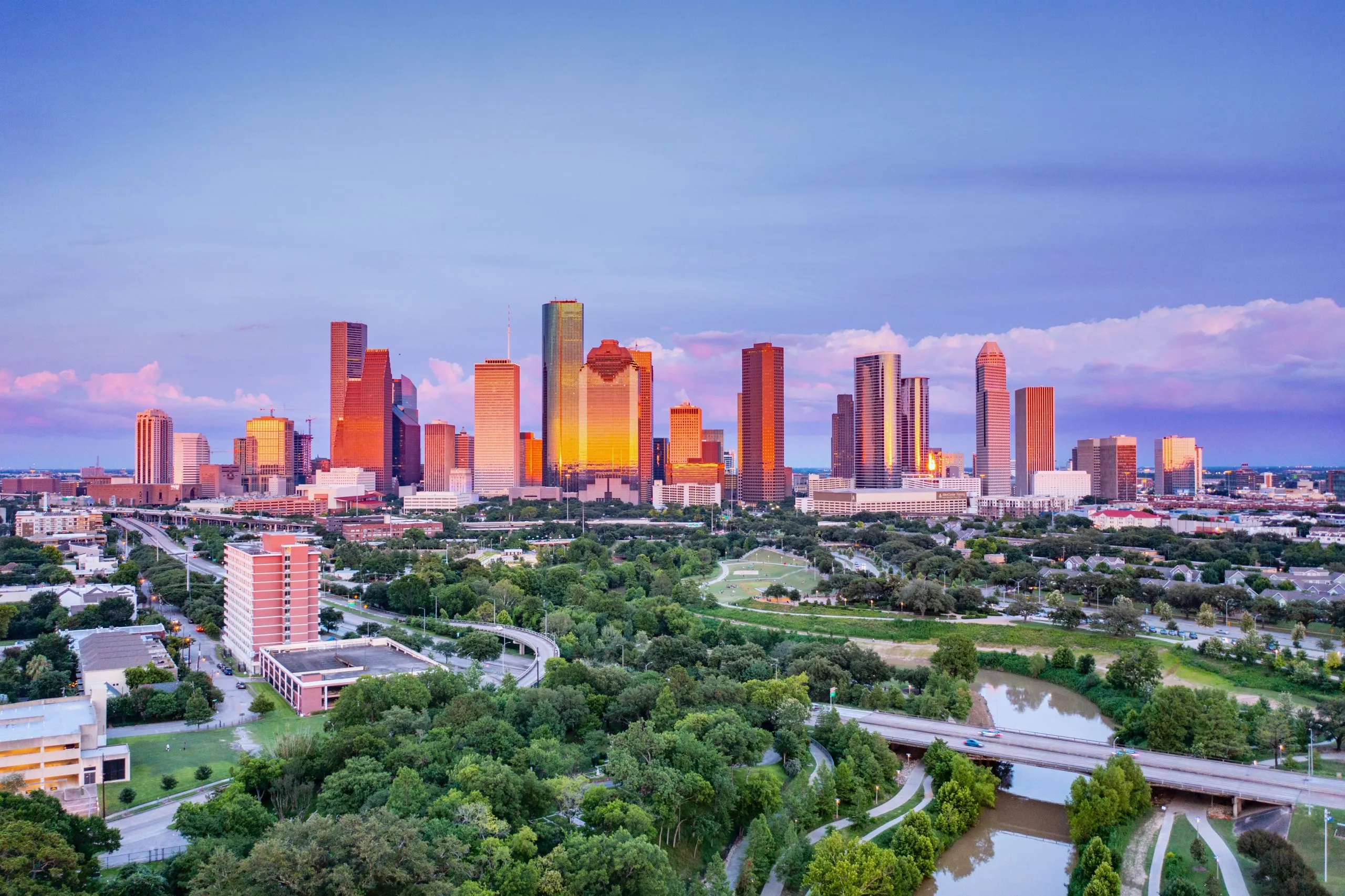 A vibrant cityscape of Houston, Texas, with skyscrapers glowing at sunset and lush greenery below.