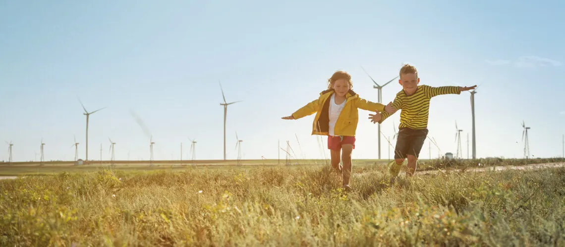 Children run joyfully in a grassy field with wind turbines in the background under a clear sky.