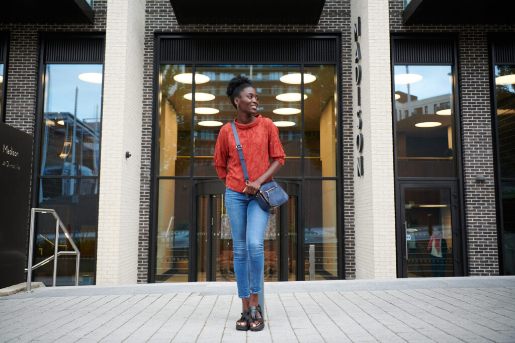 A person stands outside a modern building, wearing a red top, jeans, sandals, and a crossbody bag.
