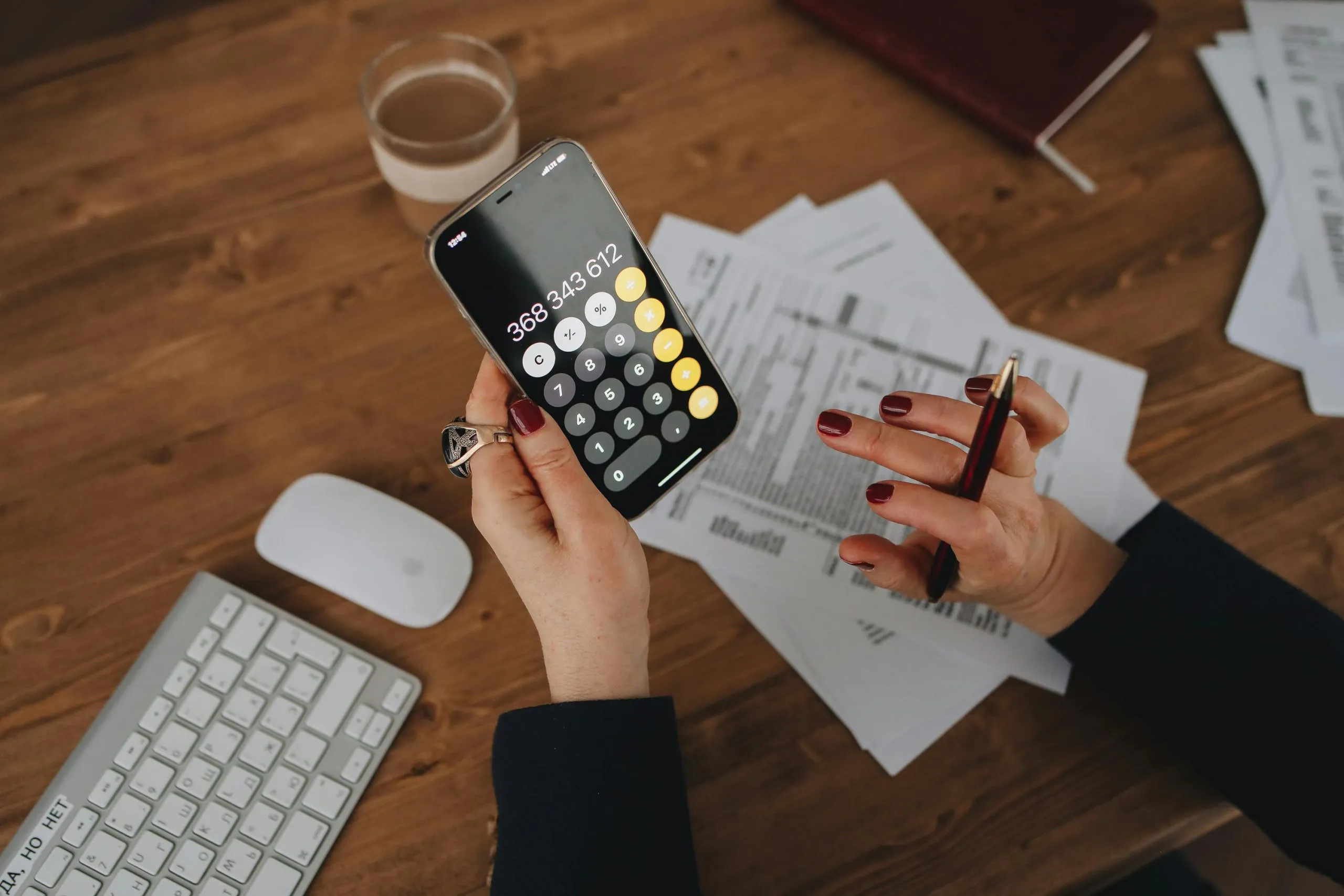 A person calculates on a smartphone while reviewing documents at a wooden desk.