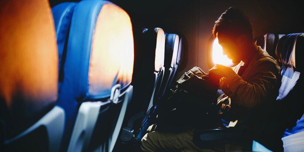 A person seated on an airplane reads under warm sunlight streaming through the window.