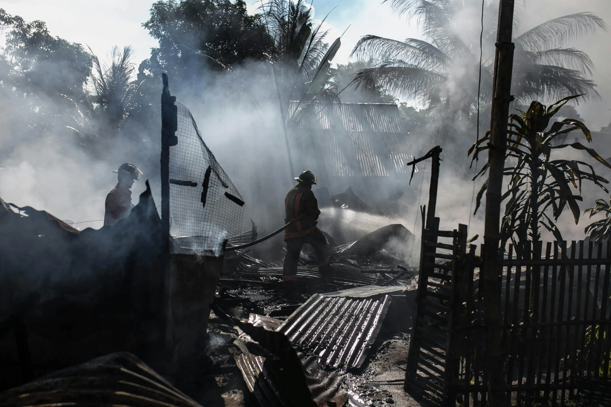 A firefighter battles smoke and flames amid the charred remains of a structure in a rural area.