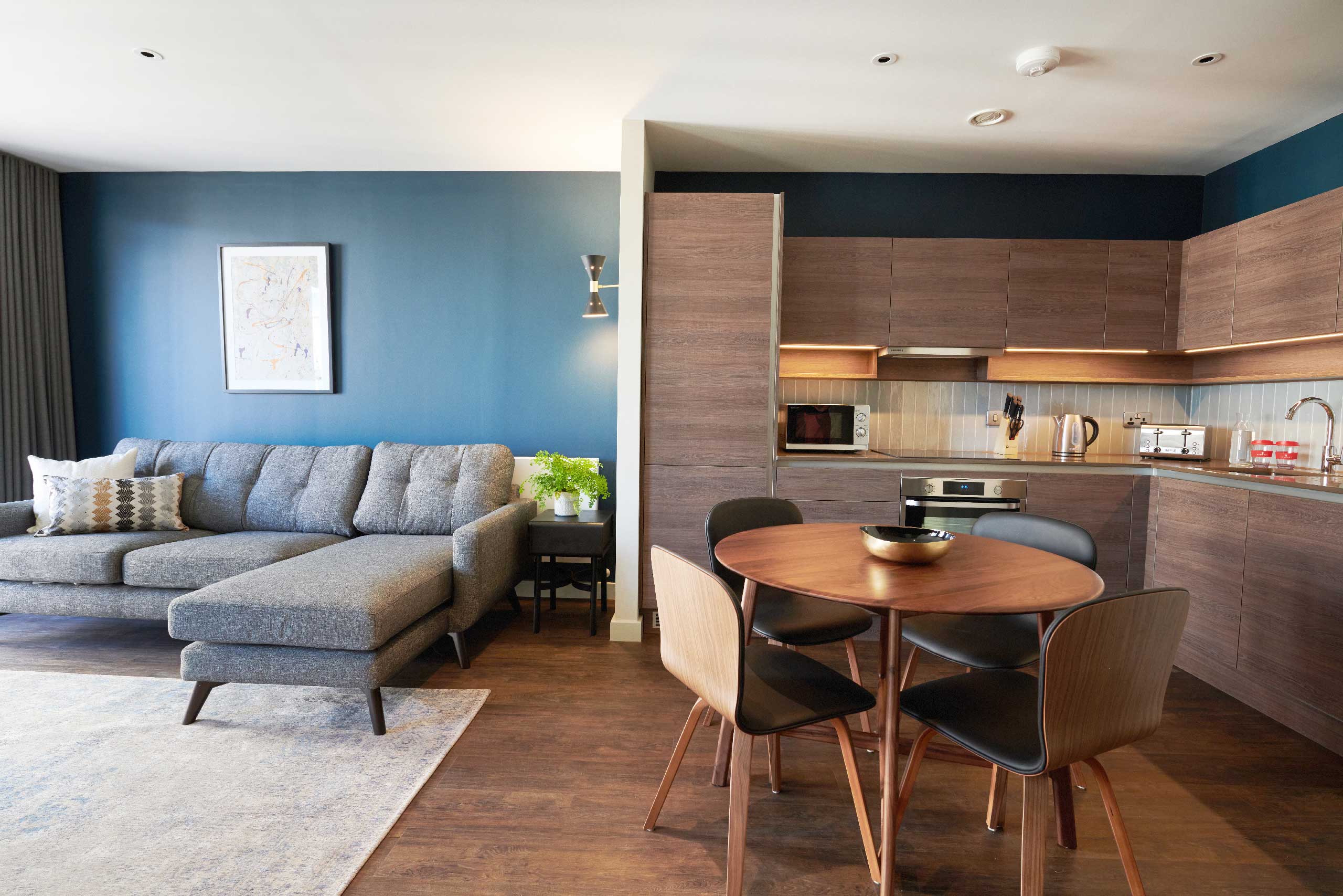 Modern open-plan living area with a blue accent wall, gray sofa, and wooden kitchen-dining setup.
