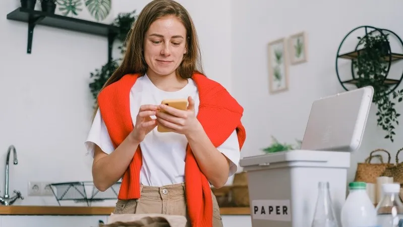 A person stands in a kitchen, holding a phone, with a recycling bin labeled "PAPER" nearby.