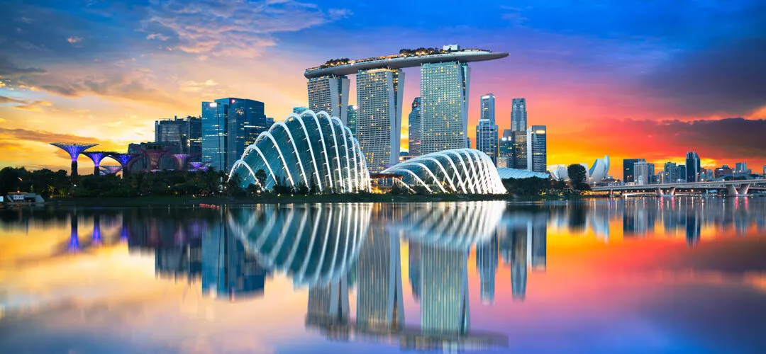 Singapore skyline at sunset, featuring Marina Bay Sands and Gardens by the Bay reflected in water.