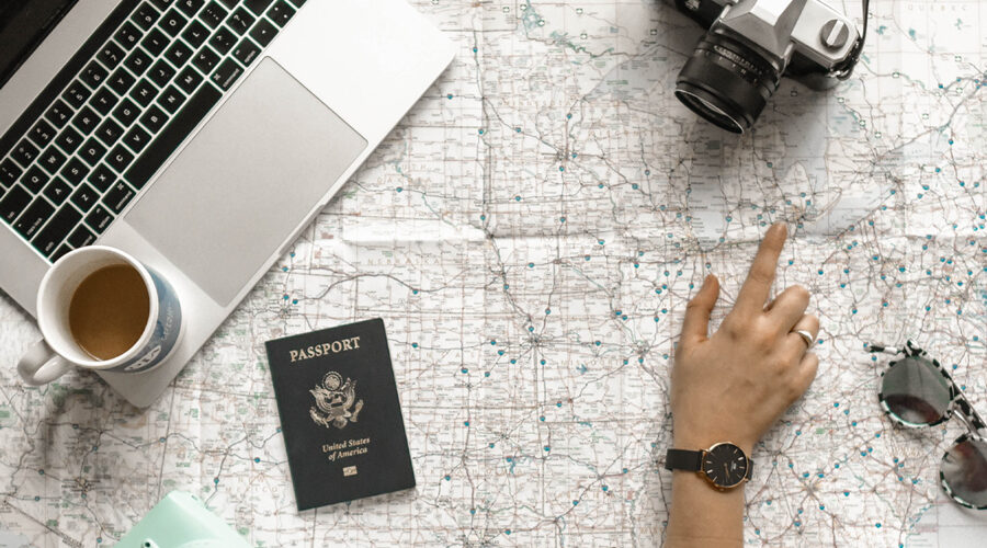 A travel planning setup with a map, laptop, passport, coffee, camera, and sunglasses.