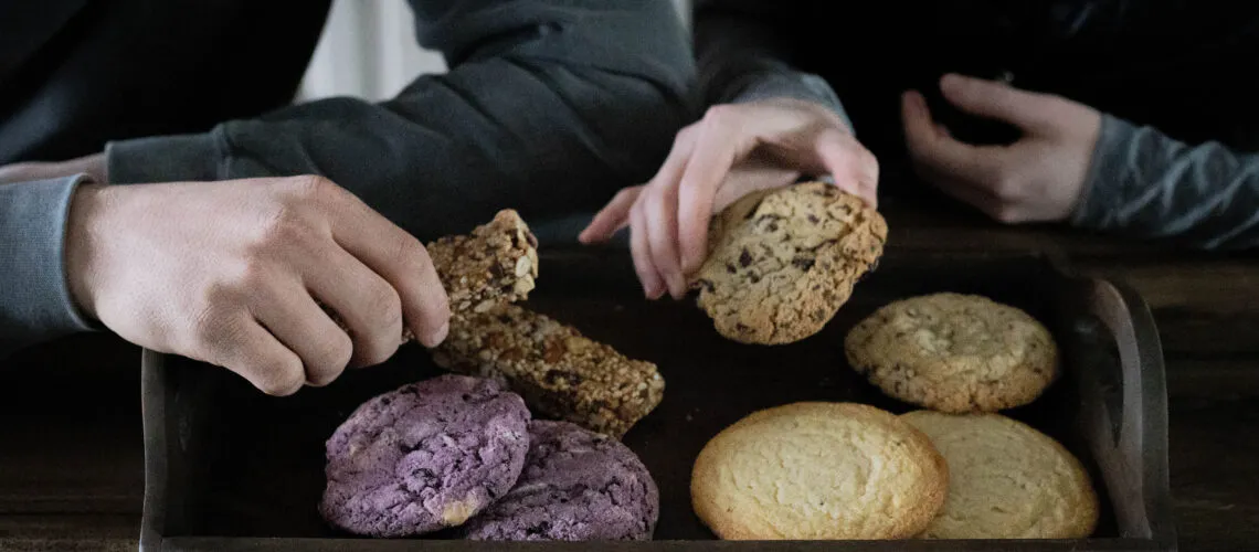 Two people reach for assorted cookies and a granola bar on a wooden tray.