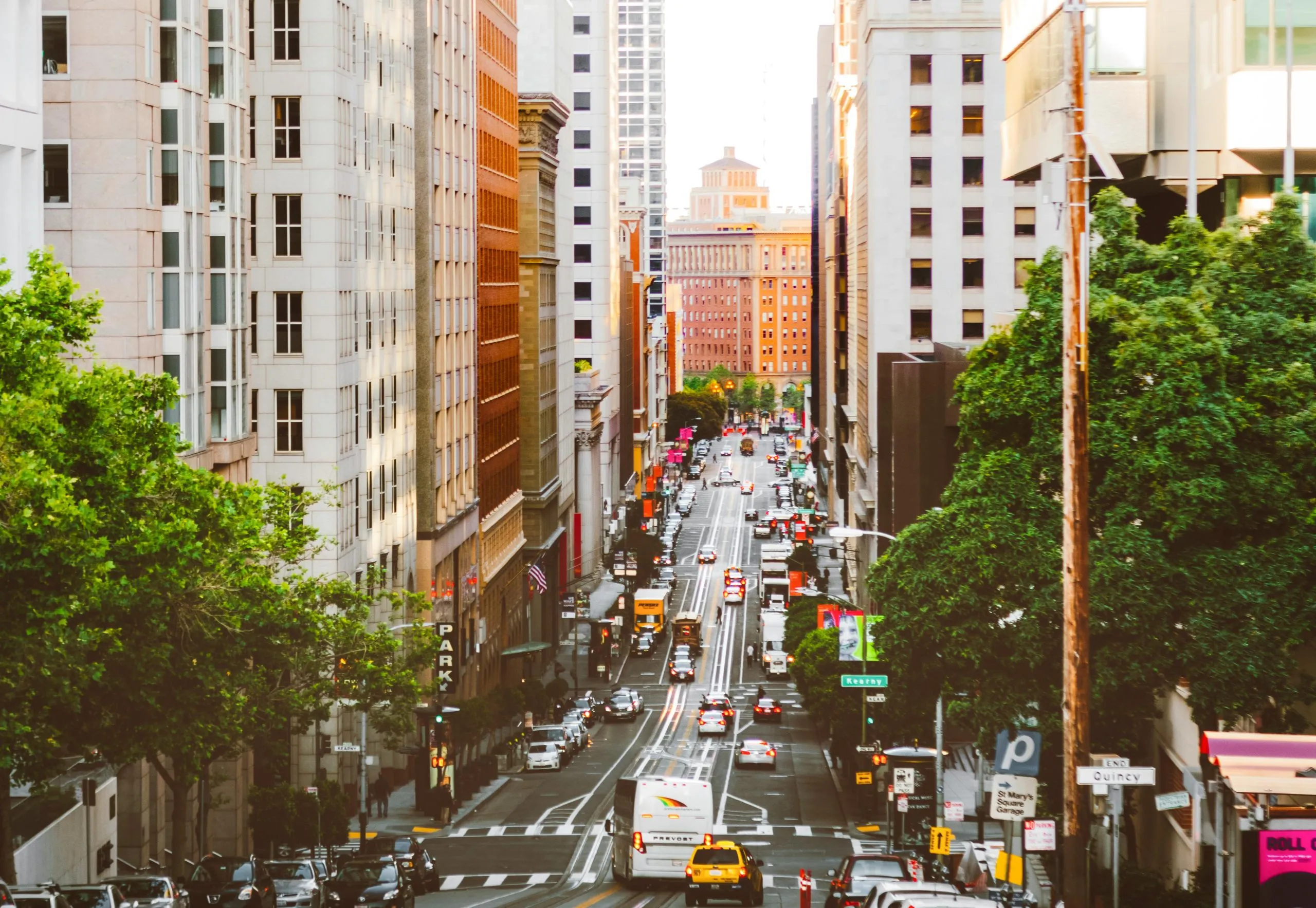 A bustling urban street lined with tall buildings, trees, and vehicles during daylight.