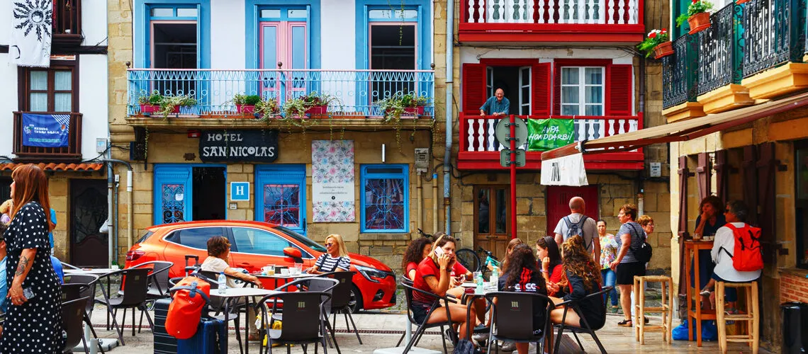 A vibrant street scene features colorful buildings, outdoor dining, and lively social interactions.