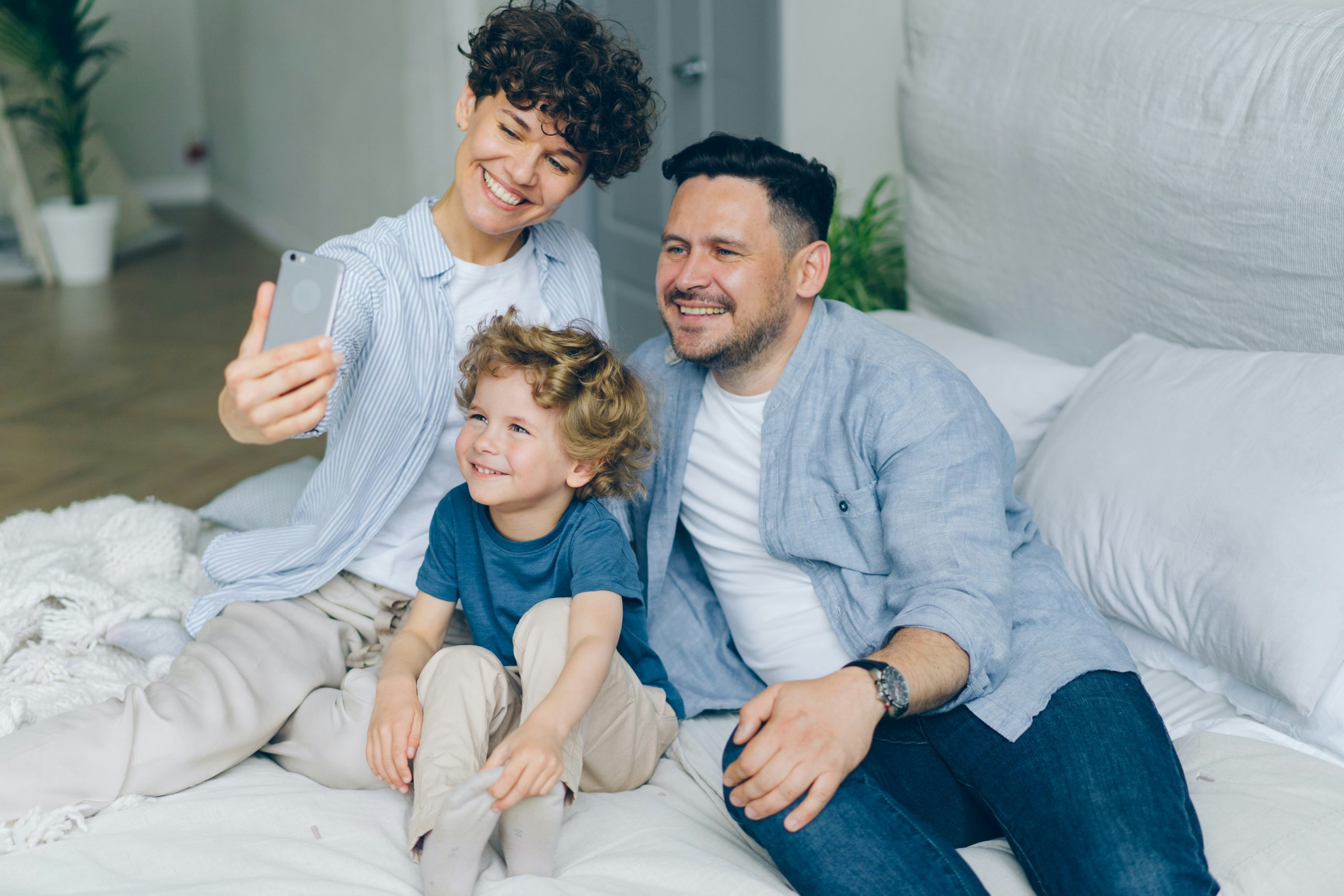 A family sits together on a bed, with one person holding a smartphone for a selfie.