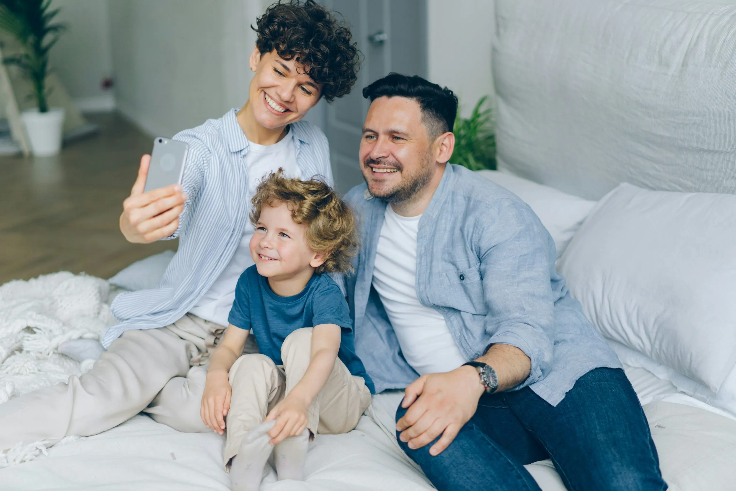 A family sits together on a bed, with one person holding a smartphone for a selfie.