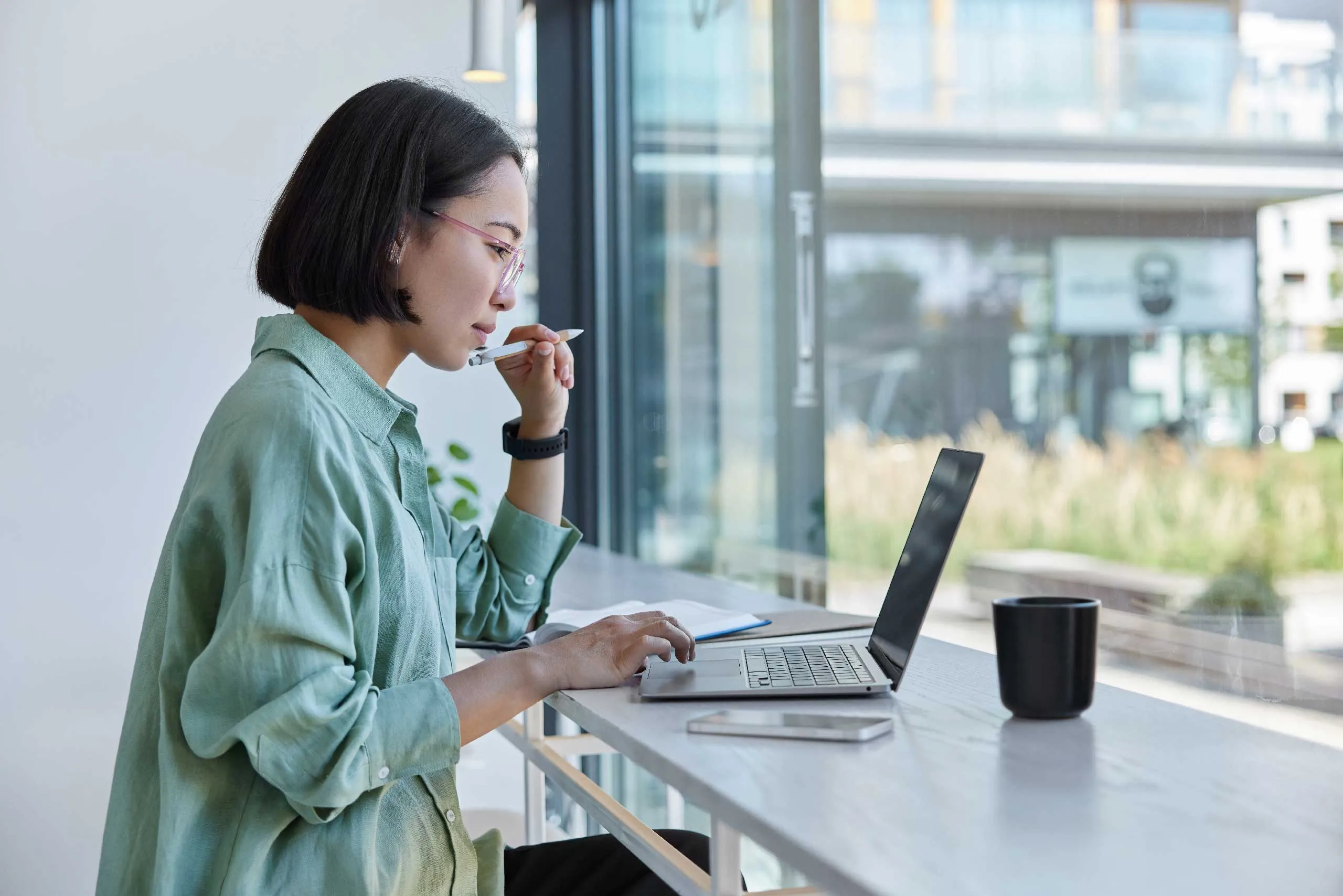 A person works on a laptop at a bright, modern workspace with a mug and smartphone nearby.