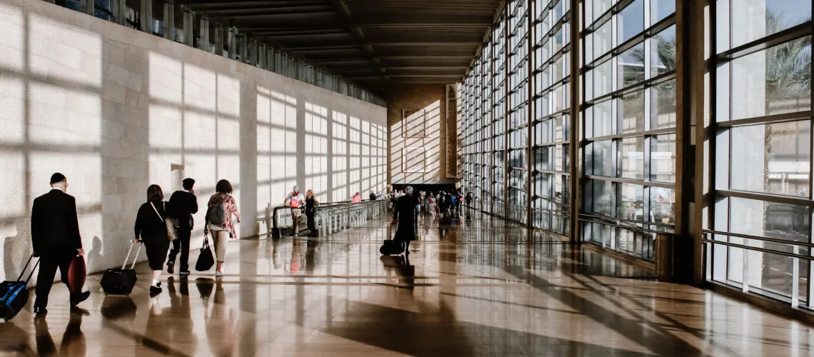 Travelers walk through a sunlit airport terminal with large glass windows and reflective flooring.