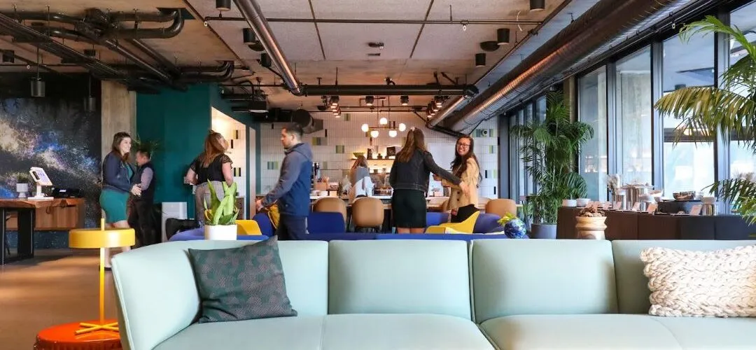 A modern, open-concept workspace with vibrant seating, greenery, and people interacting near a café area.